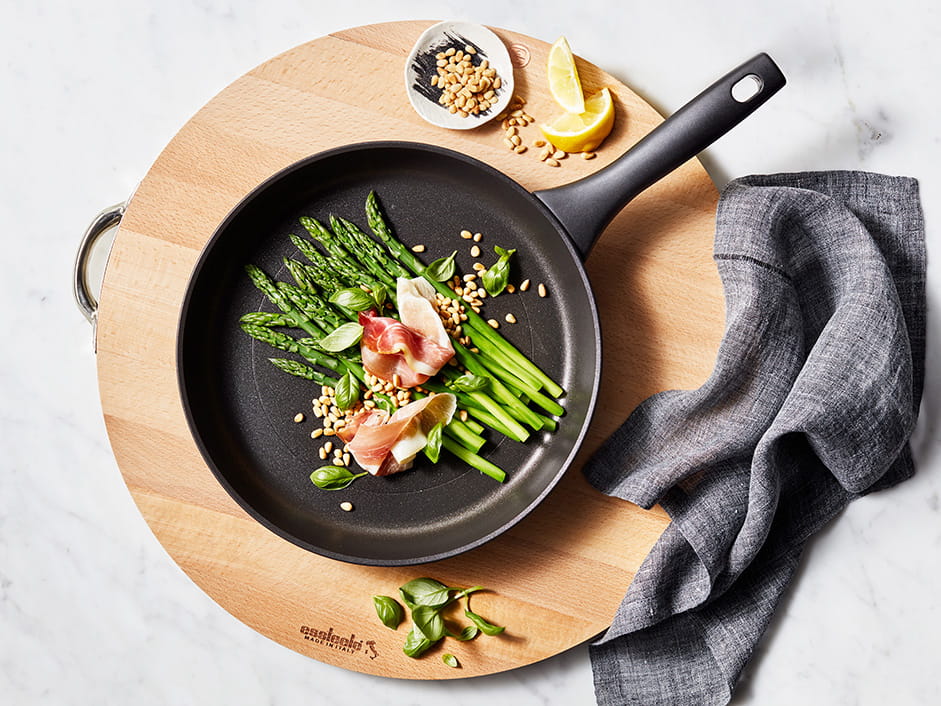 Fried asparagus with bacon in a black frying pan on a wooden board.